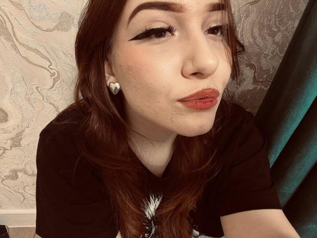 MauritaKelsch with heart earrings, pouty red lips, black eyeliner, wavy brown hair, wearing a black shirt, looking into camera, slight smile, fair skin, small nose.