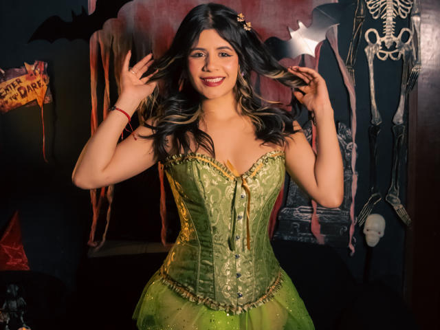 Sheyla Spooner, met donker golvend haar, een groene korset met veters en een groene tule rok, houdt haar haar vast, glimlacht en laat haar borsten zien, in een setting met een Halloween-thema..