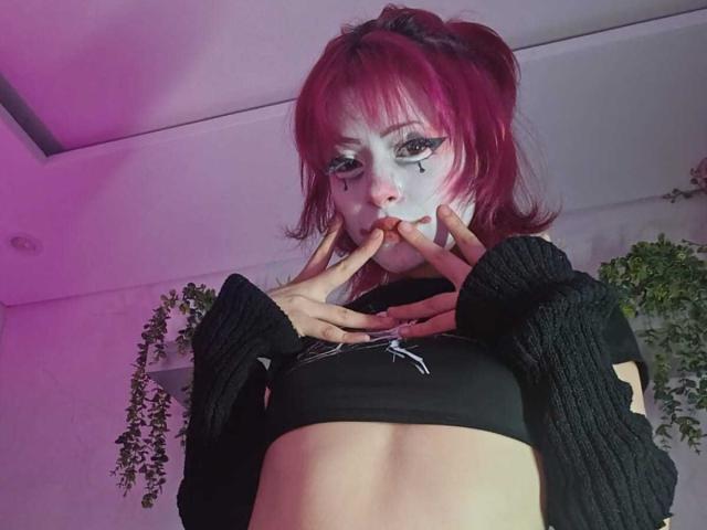 ViolettClay with pink hair, white face paint, black crop top, black ribbed sleeves, holding fingers to mouth, midriff visible, pouty expression, pink and purple lighting.