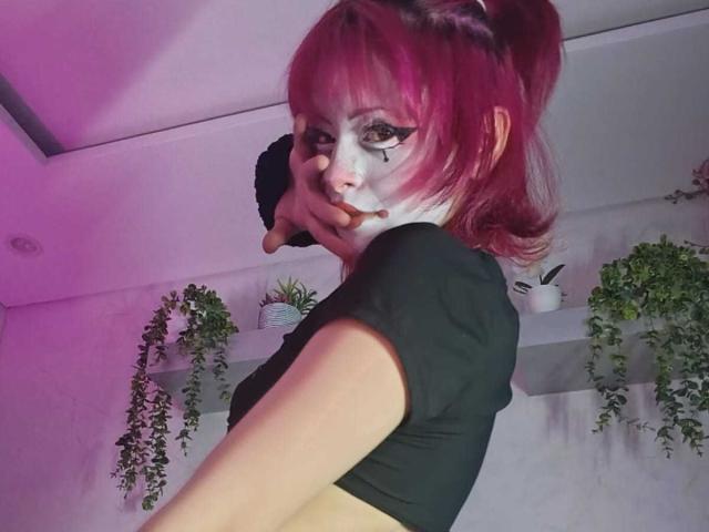 ViolettClay, com cabelo rosa, cropped top preto, maquiagem branca no rosto e delineador preto, cobrindo a boca com a mão, olhando por cima do ombro, mostrando os seios..