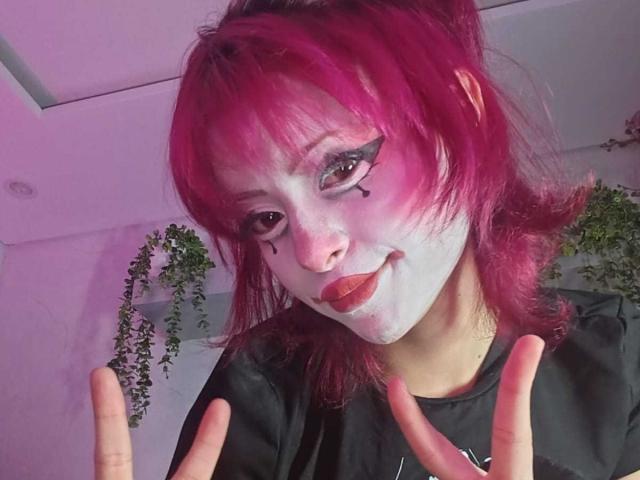 ViolettClay with pink hair, white face paint, black eyeliner, red lipstick, and a cute smile, making a playful gesture with her fingers. She's wearing a black shirt.