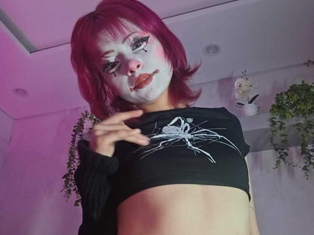 ViolettClay, com cabelo rosa, maquiagem branca no rosto e uma cropped preta que mostra parte do abdômen, com um desenho de aranha, apontando para a câmera. Suas nádegas estão parcialmente visíveis..