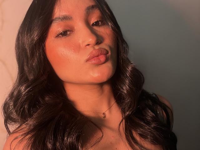 MiiaPalmer met golvend zwart haar, glanzende bruine huid, volle lippen en een subtiele ketting, die lichtelijk opzij kijkt in warm, zacht licht. Borsten niet zichtbaar..