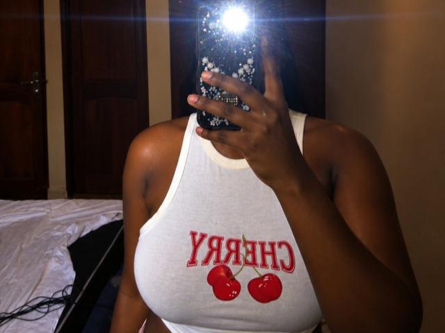 Orazl with dark skin, large boobs in a white tank top with red cherries and "YAH" text, holding a sparkly phone in a mirror selfie.