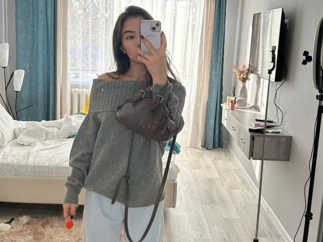 AnneKofron with long brown hair, wearing an off-shoulder grey sweater and white pants, holding a brown bag and phone, mirror selfie, bedroom with bed and wall mirror.