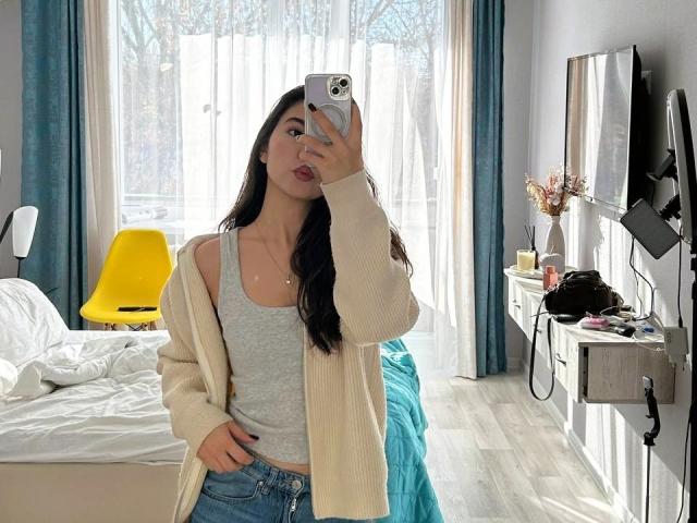 AnneKofron with long black hair, pouty lips, wearing a gray tank top, beige cardigan, and blue jeans, taking a mirror selfie, holding an iPhone, in a bright bedroom.