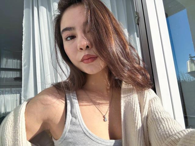 AnneKofron with medium tits, brown hair, light skin, wearing a gray tank top and white cardigan, sunlight in her face, necklace with small pendant, serious expression.
