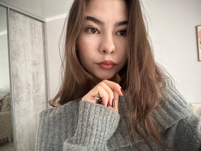 AnneKofron with long brown hair, gray sweater, and pouty lips, her finger touching her chin, showing maroon nails, looking directly at the camera.
