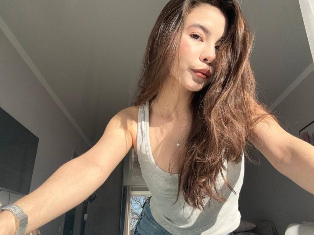 AnneKofron with long brown hair, wearing a grey tank top, showing small tits, standing in sunlit room, slightly pouting with arms out, wearing a grey watch.
