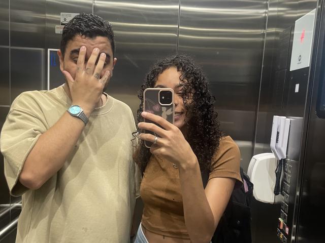 GabyAndVictor with curly brown hair taking a selfie in an elevator, wearing a brown crop top, and Victor with short dark hair, covering his face, in a beige tee.