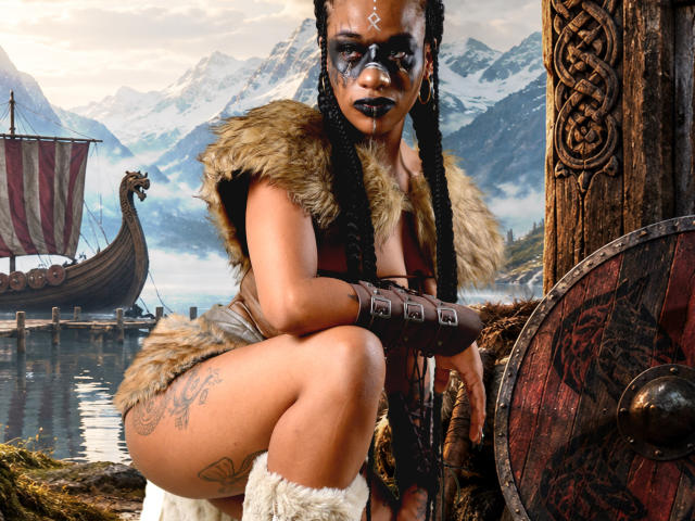 KarlaRicochet, di carnagione scura con capelli intrecciati, trucco da guerra nero, abbigliamento in pelle e pelliccia, tatuaggi sulla coscia, dalla ginocchia in su, che tiene uno scudo, con una nave vichinga sullo sfondo, montagne e acqua..