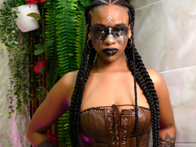KarlaRicochet avec du maquillage sombre sur le visage, du rouge à lèvres noir, des cheveux tressés, un corset marron, du maquillage noir sur le nez, un symbole blanc sur le front, des boucles d'oreilles cerceaux et de la bave sur le menton..