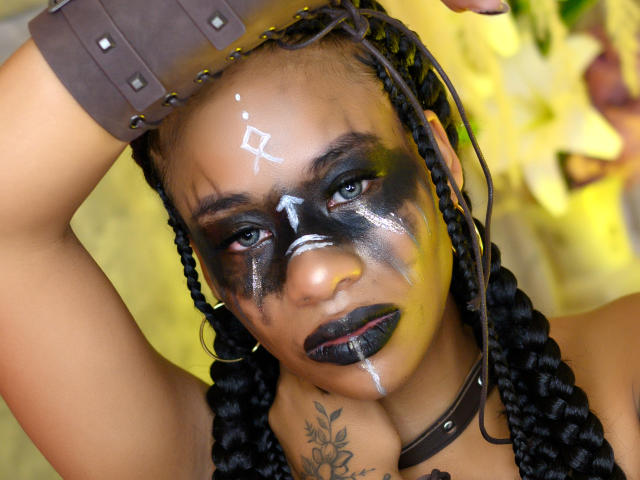 KarlaRicochet has dark braided hair, yellow and black face paint, black lipstick, a white diamond mark on her forehead, and a brown leather wrist cuff. Her tattoo is visible.
