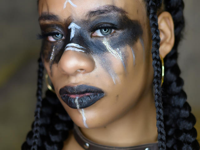 KarlaRicochet has dark makeup on her face with white streaks, black lipstick, and braided hair. She's wearing gold hoop earrings and has a serious expression.