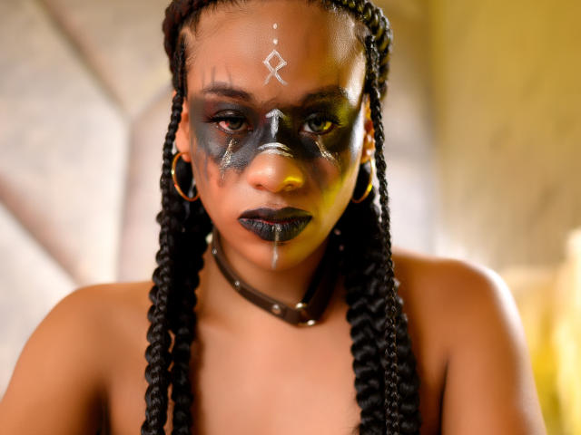 KarlaRicochet has dark braided hair, black face paint, and a white mark on her forehead. Her black lipstick drips, and she wears a brown choker and gold hoop earrings.