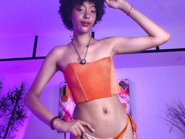 KiaraKamau with curly hair, orange mesh tube top, and orange string bikini, standing, hand on hip, purple lighting, navel piercing, confident smile, wrist bracelet, necklace.