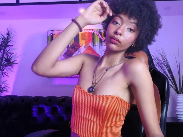 KiaraKamau with curly black hair, wearing an orange strapless dress, looking at the camera, hand near head, purple and pink lighting, black chair, and plant in background.