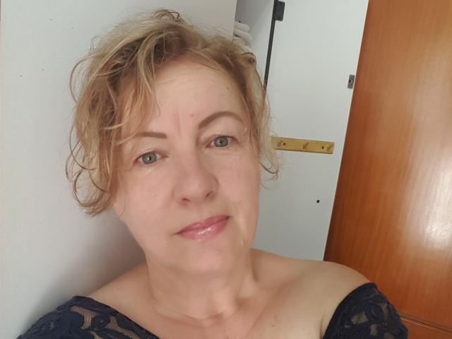 LillaBelle has short, messy blonde hair, light green eyes, and a slight smile. She's wearing a black lace top, showing her bare shoulders and neck. She's in a doorway.