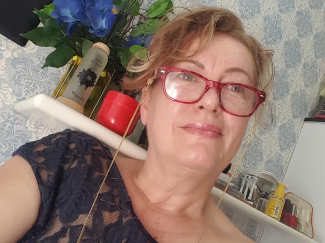 LillaBelle, avec ses courts cheveux blonds en bataille, ses lunettes rouges et son haut en dentelle, a une peau brillante et une expression légèrement hautaine. Une bougie rouge et des fleurs bleues sont visibles en arrière-plan..