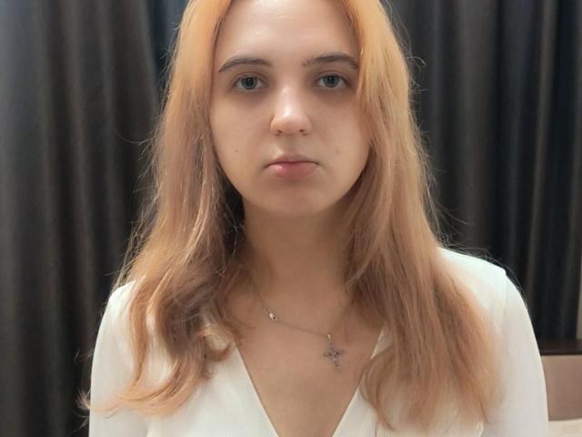 JonieAamot tiene el cabello castaño claro, de largo hasta los hombros, ojos azules y una expresión neutra. Lleva una blusa blanca con un collar pequeño, y muestra un rostro ligeramente serio..