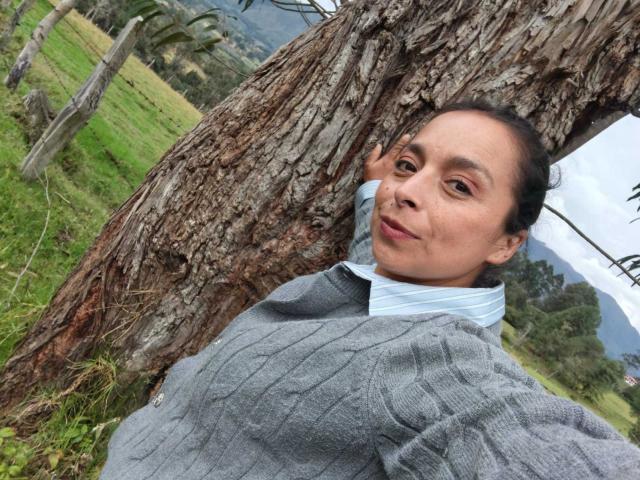 StangelarRivera, con un seno di media grandezza, indossa un maglione grigio a coste e una camicia blu a righe, appoggiata a un albero, sorridente, con erba verde e montagne sullo sfondo, capelli scuri..