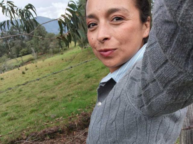 StangelarRivera with medium brown skin, dark brown eyes, and light pink lipstick, wearing a gray cardigan over a blue shirt, raising her arm, outside in a grassy field.