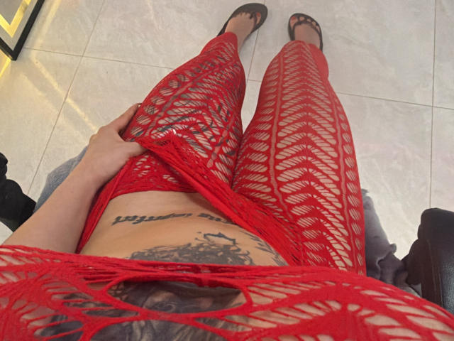 As leggings de rede vermelhas com padrão de treliça da MiaValenttine, que revelam sua barriga tatuada e sua vulva, com a mão dela puxando o tecido. Ela está usando sapatilhas pretas, mostrando os dedos dos pés..