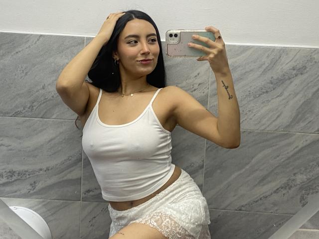 CarlaFranco with long black hair, white tank top showing her small tits, white lace shorts, taking a mirror selfie, hand in hair, tattoo on right arm, smiling.