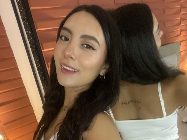 CarlaFranco with long black hair, white tank top, small gold earrings, nose ring, smiling, back tattoo visible in mirror, soft pink lipstick, light brown eyes, medium skin.
