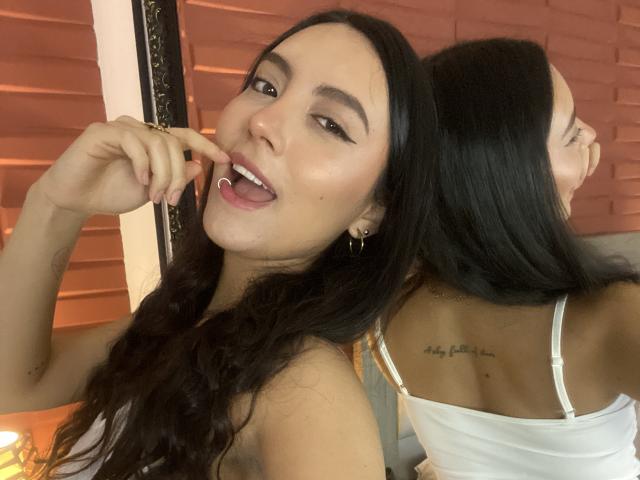 CarlaFranco with long black hair, tongue out, finger on lip, looking at camera, white strap top, tattoo on back, reflection in mirror, golden earrings, sexy, confident.