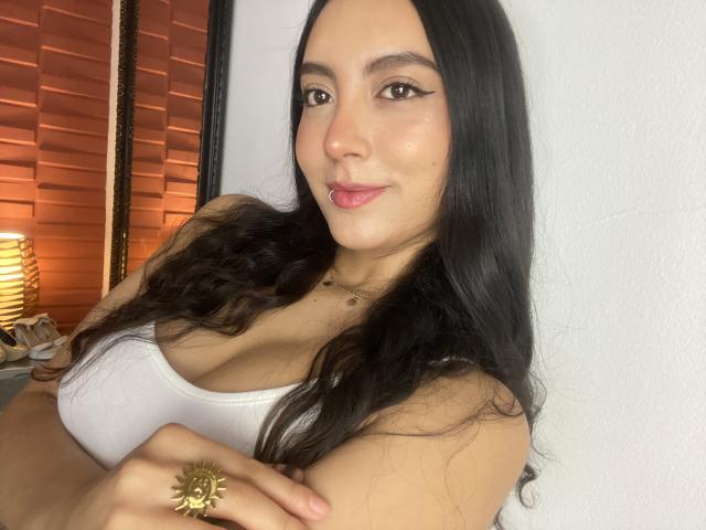 CarlaFranco with long black hair, wearing a white tank top, showing her tits, has a sun ring on her right hand, and a subtle lip piercing, smiling.