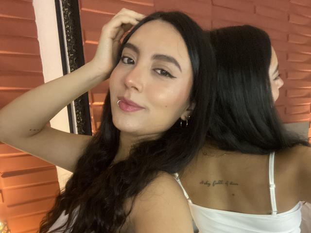 CarlaFranco with long black hair, brown eyes, and pink lips, wearing a white top, showing a tattoo on her back, and a reflection of her head in a mirror.