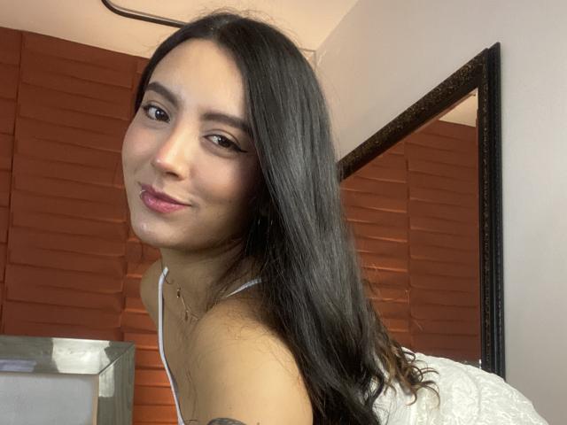 CarlaFranco with long black hair, light skin, and a small nose ring, smiling at the camera, wearing a white tank top, showing her tits in the selfie.
