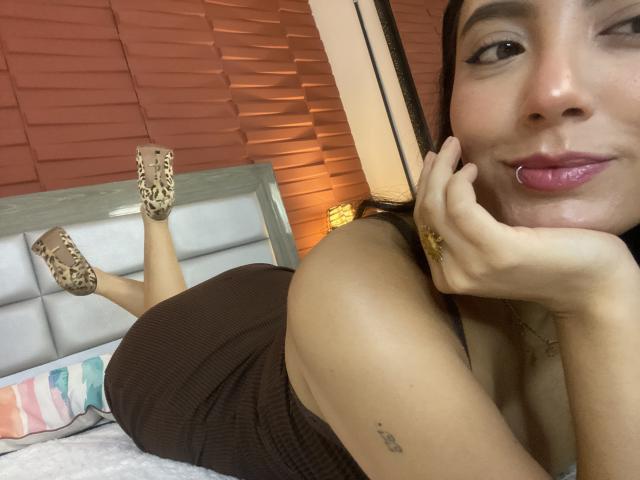 CarlaFranco lying on bed, winking, with her head resting on hand, brown dress, leopard print heels, pierced lip, small tattoo on arm, brown skin, smiling, legs up.