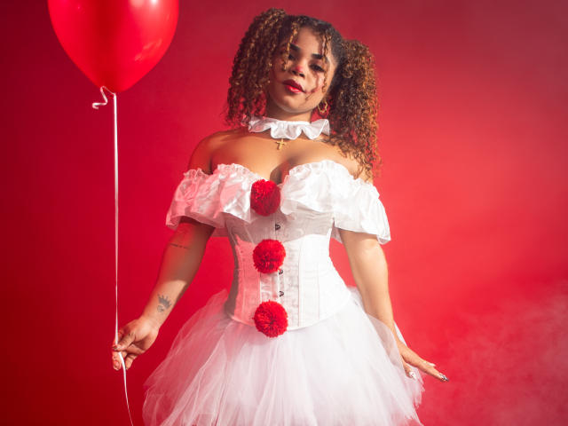 NahiaraReina with curly hair, wearing a white off-shoulder dress with red pom-poms, holding a red balloon, has medium tits, and a tattoo on her right arm.