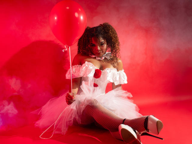 NahiaraReina with curly hair, white off-shoulder dress, fishnet stockings, holding red balloon, sitting, red high heels, fluffy white skirt, red background, looking seductively at camera.