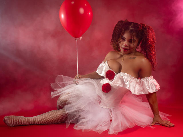 NahiaraReina with curly red hair, white off-shoulder dress, fishnet stockings, holding red balloon, sitting on red floor, white tulle skirt, large tits, red pom-poms, smiling, red light.