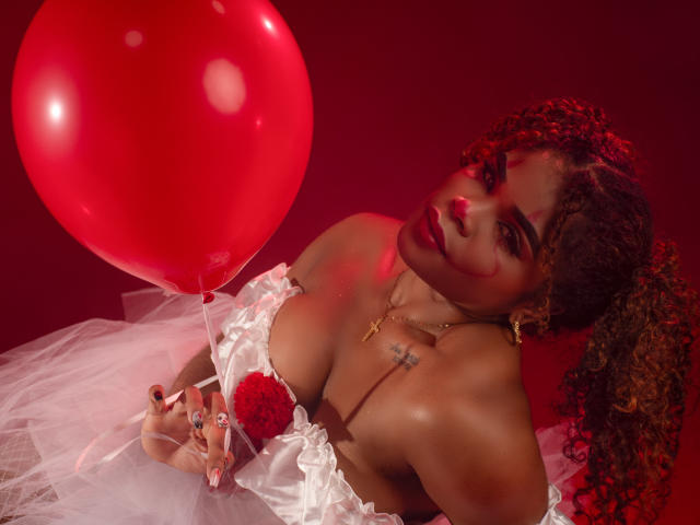 NahiaraReina with curly hair, red lipstick, white off-shoulder top, holding red balloon, white tulle, and rose, gold necklace, small tattoo, looking up, red light. Boobs, tats, rose.