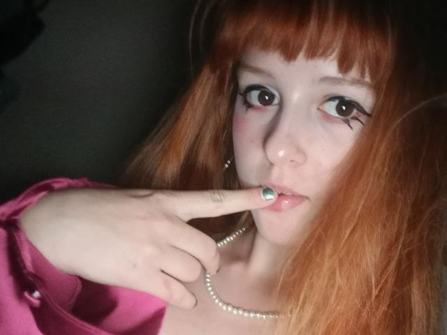 LouieOrner with red hair, pearl necklace, and pink shirt, has finger in mouth, showing green nail polish, and has black eyeliner on her large eyes.