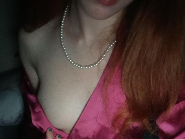 LouieOrner's tits are visible with pink satin and a pearl necklace, showing fair skin and red hair, with dark green painted nails.