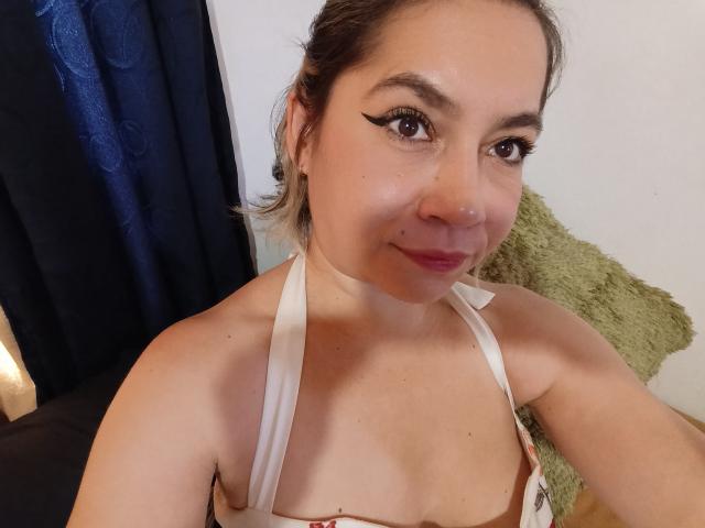 MsJustineBeaux with dark eyebrows, eyeliner, nose ring, and light brown hair in a bun, smiling, wearing a white halter top, showing small tits, in a room.