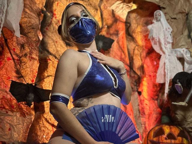 LeylaJamesX mit blauem, glänzendem Oberteil, passender Maske und Armbändern, hält einen Fächer, zeigt Tattoos, hat eine mittlere Brustgröße, trägt ein Halloween-Kostüm, steht mit einer Hand auf der Brust, in einer gruseligen Umgebung..