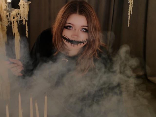 YoureGift, com cabelos castanhos desalinhados, maquiagem com um sorriso costurado em preto, rosto esfumaçado e um robe escuro, segurando velas, cercada por fumaça densa. Os olhos de YoureGift estão arregalados e ligeiramente assustadores..