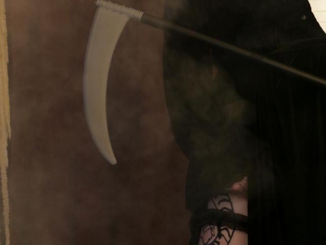 YoureGift holding a curved scythe, dressed in black with lace on their thigh, partially visible through a smoky, reflective surface.
