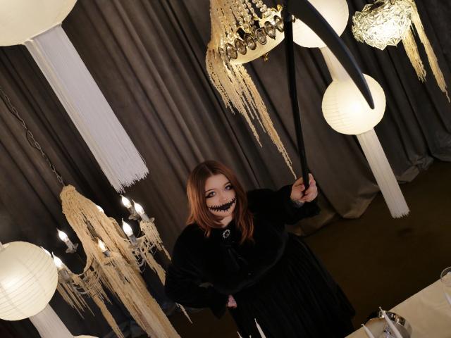 YoureGift with red hair, black dress, and toothy grin holding black ribbon, standing under ornate hanging lamps with white and gold paper decorations. Boobs, ass not visible.