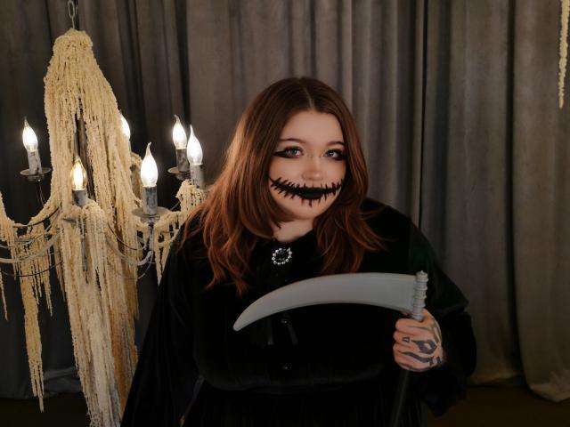 YoureGift with long brown hair, black makeup, and a toothy black grin, holding a scythe, wearing a black cloak, standing in front of a chandelier with hanging beads.