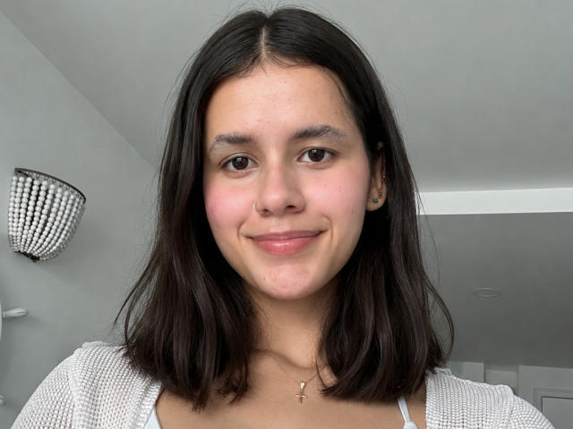 SofiaDavis1 tem a pele marrom média, cabelo preto na altura dos ombros, olhos castanhos, nariz pequeno e um sorriso discreto. Ela está vestindo uma blusa branca de tricô com um colar de cruz..