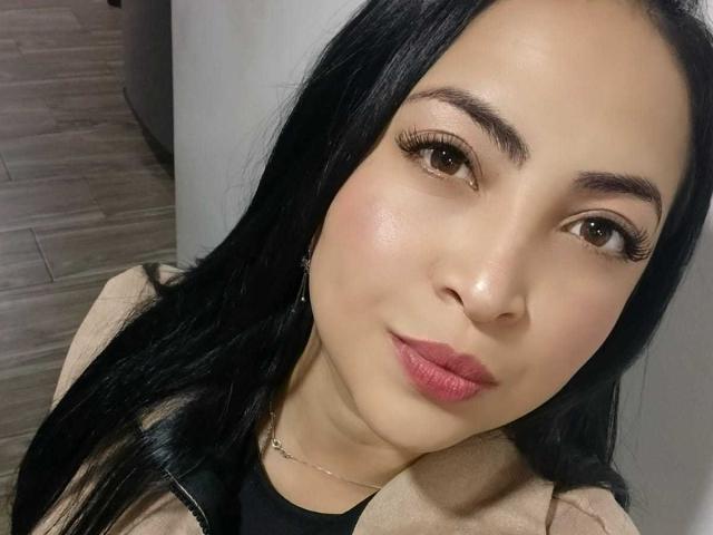 AlanaWest has a cute face with long black hair, brown eyes, and pink lipstick. She's wearing a beige jacket over a black top. Her tits are small and her pussy's not visible.