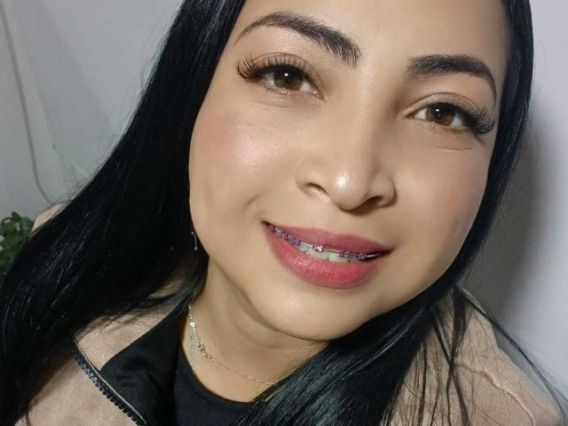 AlanaWest has long black hair, brown eyes, and pink lipstick with metallic purple braces. She's smiling with a light blush, wearing a black top and a silver necklace.