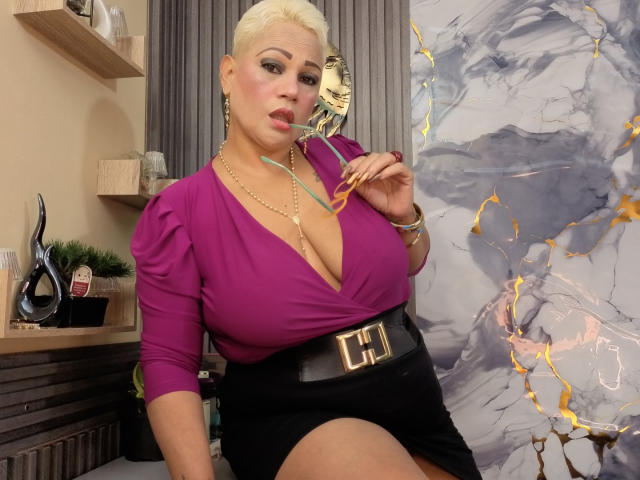 GianellaCox, avec ses cheveux blonds, son haut violet à décolleté plongeant, sa jupe noire, son collier en or et sa ceinture, tenant des lunettes, avec de grandes seins, des ongles jaunes et une expression confiante, à l'intérieur..
